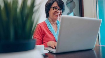 Sixty year old female teacher wearing headphones having online class via video chat on laptop computer. She is sitting on a wooden modern desk at home. Smiling and enjoying communication