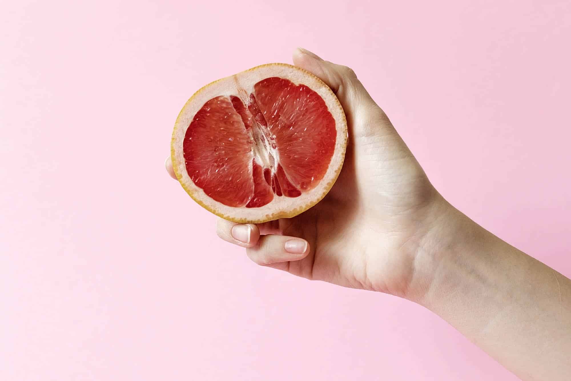 Female hands holding half grapefruit on pink background, female masturbation concept
