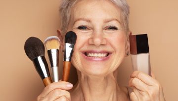 Charming elderly woman holds makeup brushes and foundation in her hands.