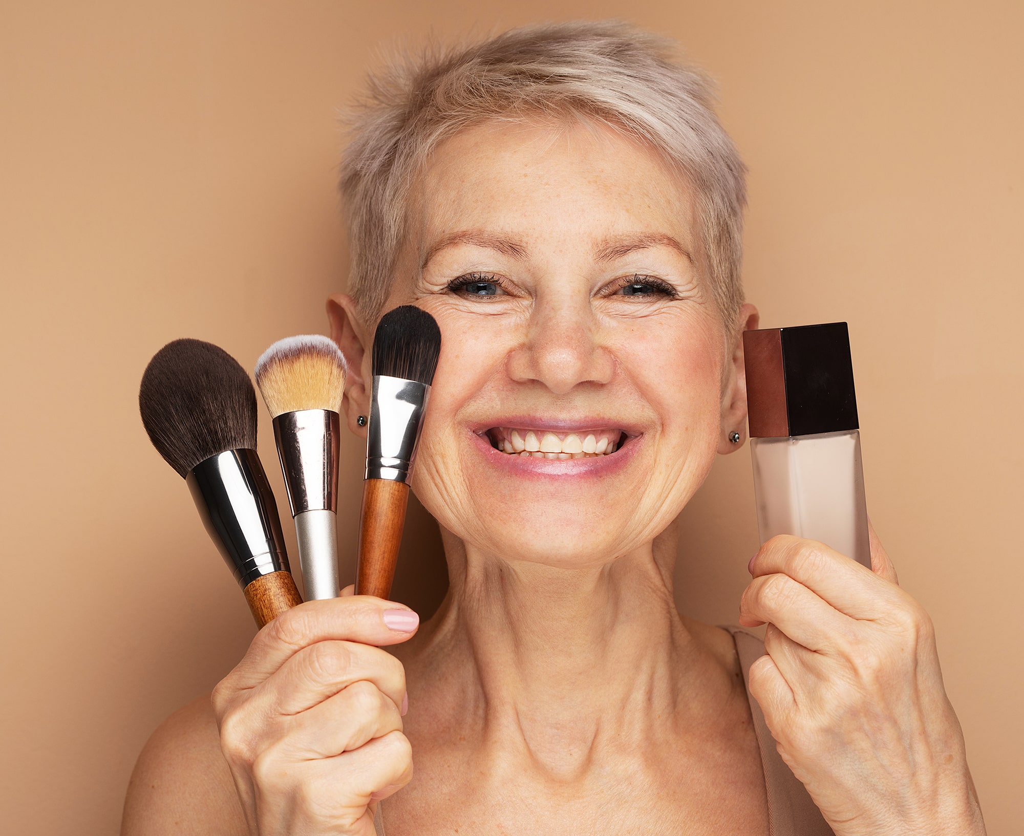 Charming elderly woman holds makeup brushes and foundation in her hands.