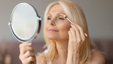 Happy senior european woman applying eyeshadow with brush, looking in mirror and smiling, female doing nude makeup