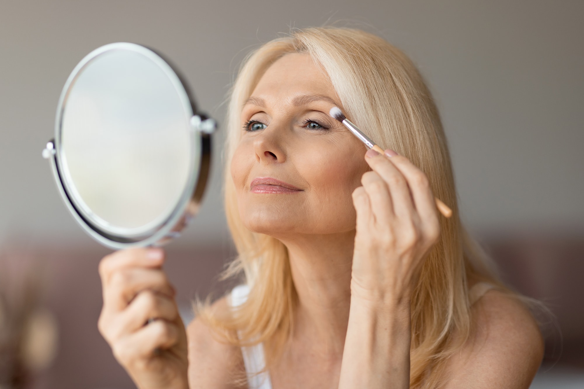Happy senior european woman applying eyeshadow with brush, looking in mirror and smiling, female doing nude makeup