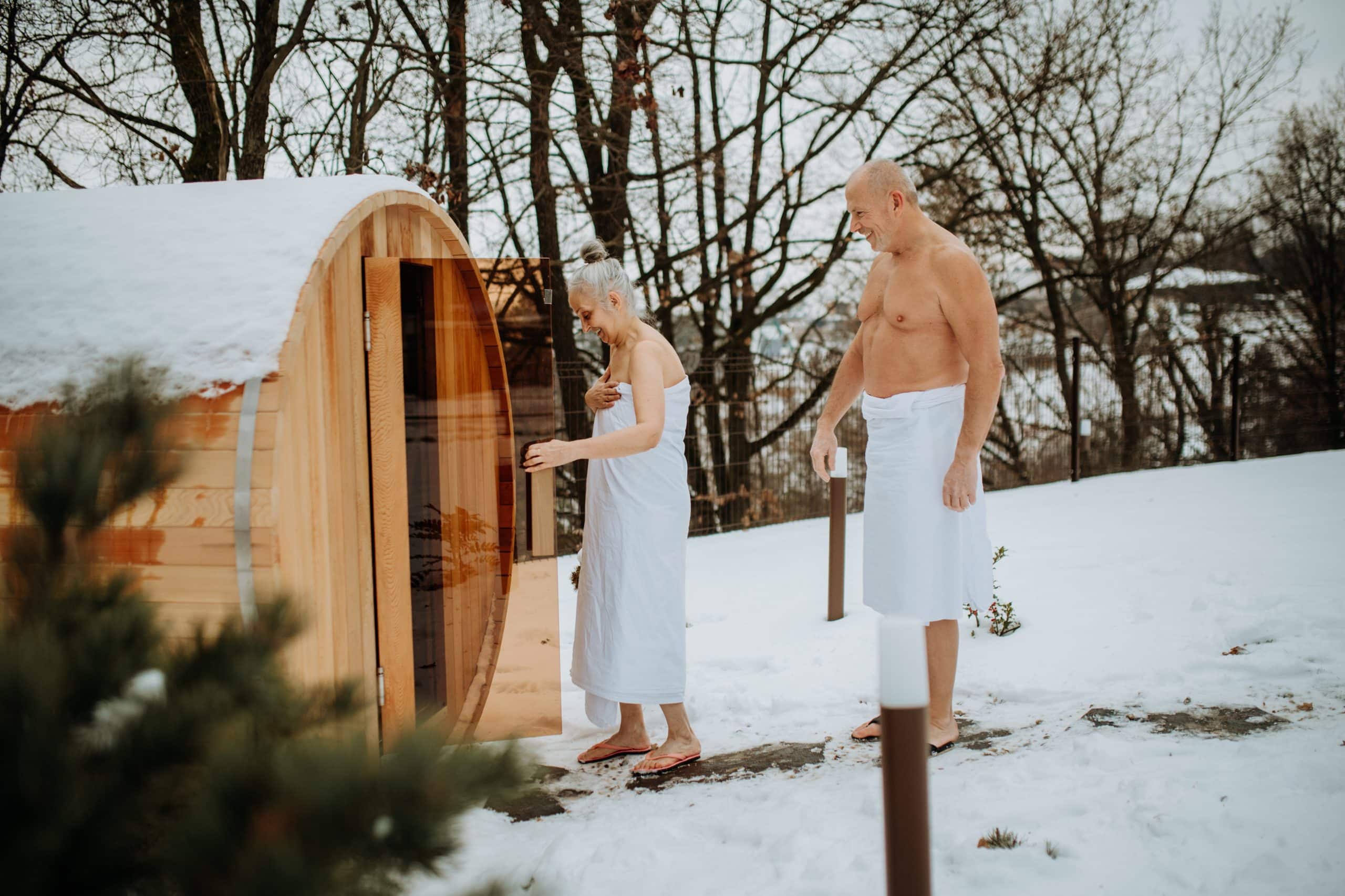 Senior woman in towel with her husband entering to outdoor sauna during cold winter day.
